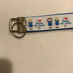 May include: A white and blue key chain with a silver key ring. The key chain has a repeating pattern of a girl scout in a blue uniform with a red heart and the text "I love Daisy Girl Scouts".