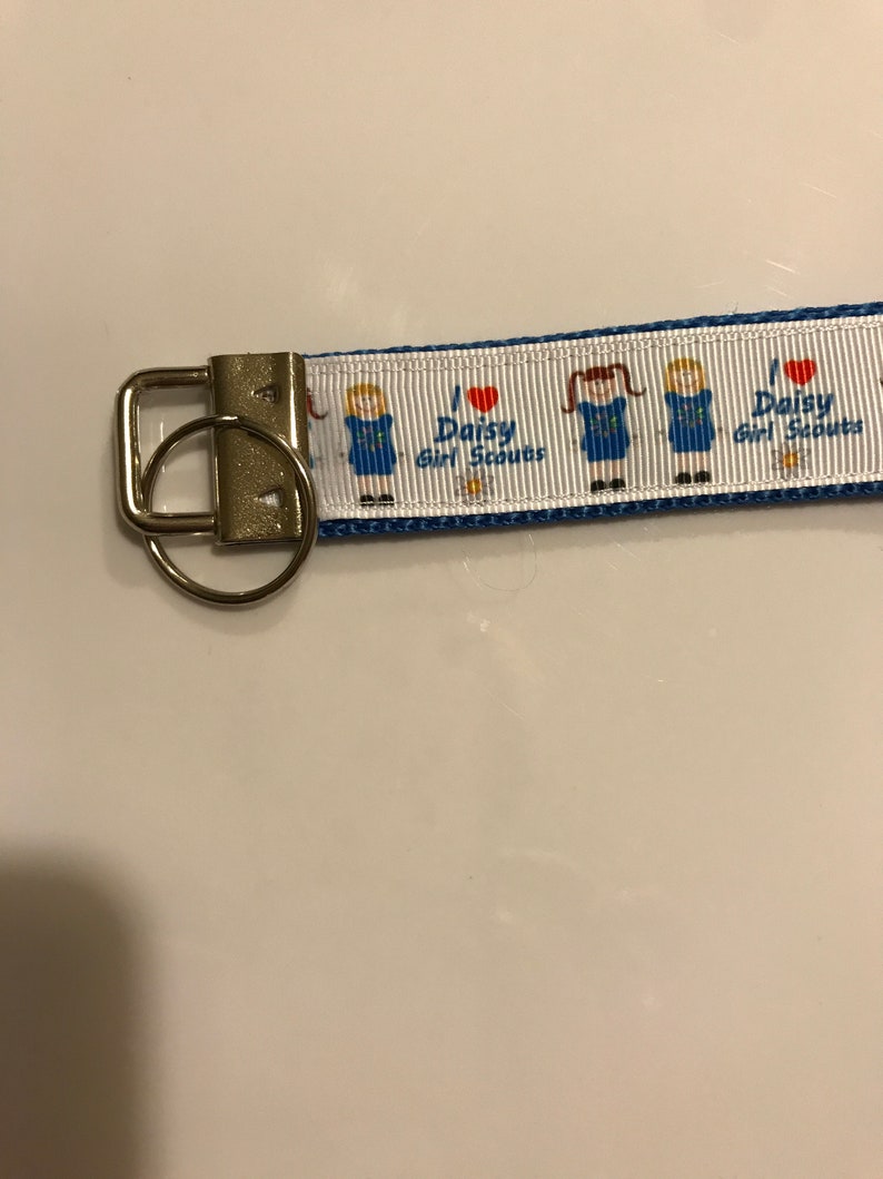 May include: A white and blue key chain with a silver key ring. The key chain has a repeating pattern of a girl scout in a blue uniform with a red heart and the text "I love Daisy Girl Scouts".