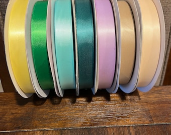 May include: Seven spools of satin ribbon in various colors, including yellow, green, light blue, teal, lavender, beige, and cream. The spools are arranged in a row on a wooden surface.