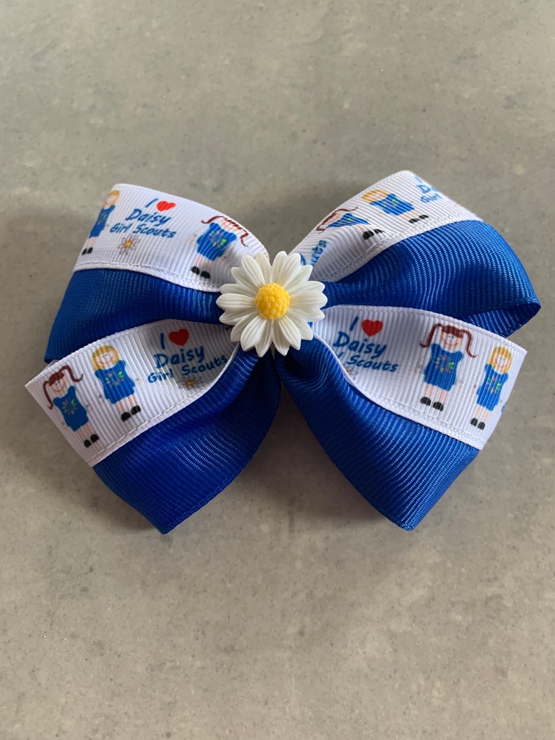 May include: Blue and white ribbon bow with a white daisy flower accent. The ribbon has a repeated pattern of a girl scout in uniform with the text "I love Daisy Girl Scouts".
