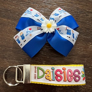 May include: A blue and white ribbon bow with a white daisy in the center. The ribbon has the text "I love Daisy Girl Scouts" printed on it. A yellow and white key chain with the text "Girl Scout Daisies" printed on it.