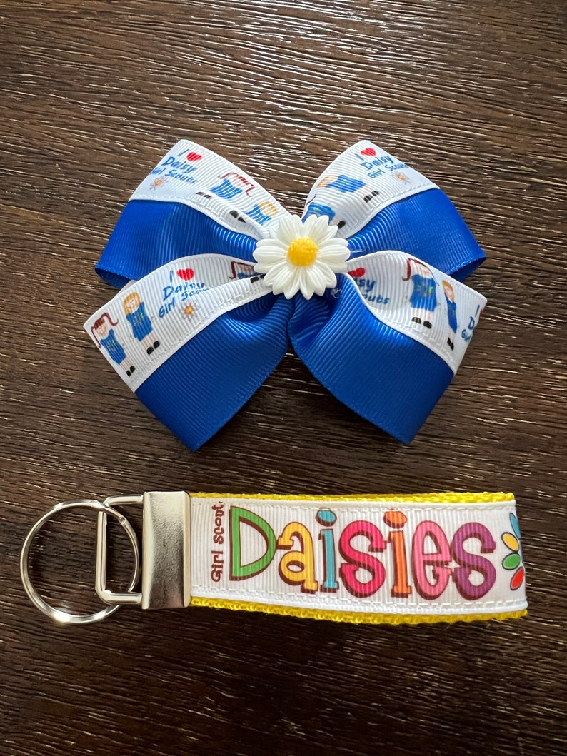 May include: A blue and white ribbon bow with a white daisy in the center. The ribbon has the text "I love Daisy Girl Scouts" printed on it. A yellow and white key chain with the text "Girl Scout Daisies" printed on it.