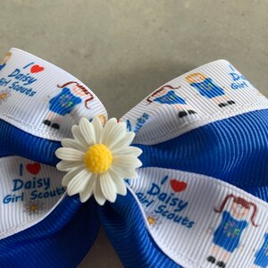 May include: A blue and white ribbon bow with a white daisy in the center. The ribbon has the text "I love Daisy Girl Scouts" printed on it.