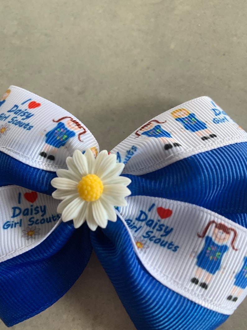 May include: A blue and white ribbon bow with a white daisy in the center. The ribbon has the text "I love Daisy Girl Scouts" printed on it.