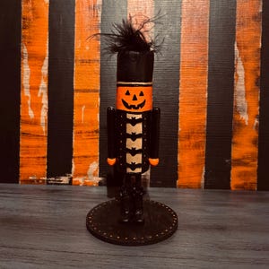 Może przedstawiać: Czarno-pomarańczowy świąteczny łamacz orzechów z motywem Halloween, z twarzą dyni, skrzydłami nietoperza i czarnym kapeluszem. Łamacz orzechów stoi na czarnej podstawie z białymi kropkami.