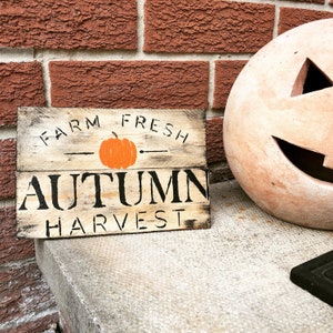 May include: A wooden sign with the text "Farm Fresh Autumn Harvest" in black lettering. A small orange pumpkin is painted on the sign.