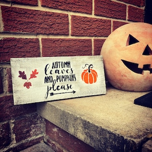 May include: A white wooden sign with black text that reads "Autumn leaves and pumpkins please" with a black arrow pointing to the right. A small orange pumpkin with a green stem is to the right of the text. The sign is sitting on a brick wall next to a large orange pumpkin with a carved jack-o'-lantern face.