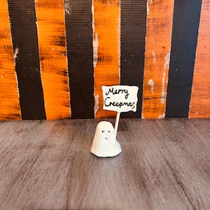 Könnte beinhalten: Eine kleine weiße Geisterfigur, die ein Schild mit der Aufschrift "Merry Creepmas" vor einem schwarz-orange gestreiften Hintergrund hält.