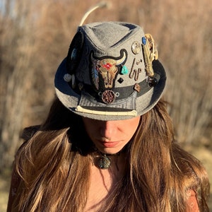 May include: A gray fedora hat with a variety of embellishments, including a bull's head design, feathers, and beads.