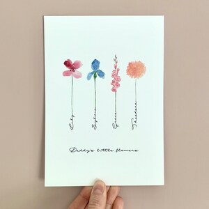 May include: A white card with a watercolor illustration of four flowers, each with a different color and name written underneath. The flowers are pink, blue, pink, and orange. The text at the bottom reads "Daddy's little flowers."