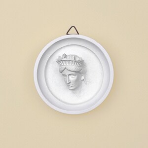 May include: A white, circular wall hanging featuring a bas-relief sculpture of a woman's face wearing a crown. The sculpture is set within a recessed circular frame. A small, dark metal hanger is attached at the top.