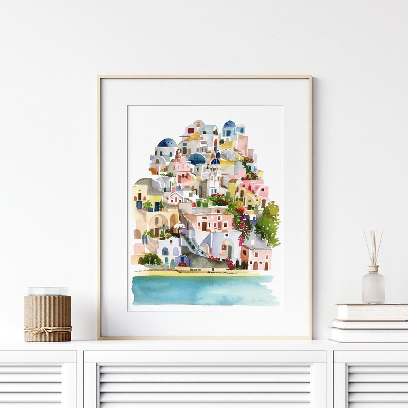 Peut inclure: Peinture &agrave; l'aquarelle d'un village blanc et bleu avec une mer turquoise au premier plan. Le village est construit &agrave; flanc de colline et poss&egrave;de de nombreuses maisons color&eacute;es avec des d&ocirc;mes bleus. La peinture est encadr&eacute;e dans un cadre en bois clair.