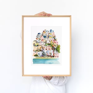 Peut inclure: Une peinture &agrave; l'aquarelle d'un village color&eacute; avec des b&acirc;timents aux d&ocirc;mes bleus, des murs blanchis &agrave; la chaux et une mer bleu turquoise au premier plan. La peinture est encadr&eacute;e dans un cadre en bois clair.