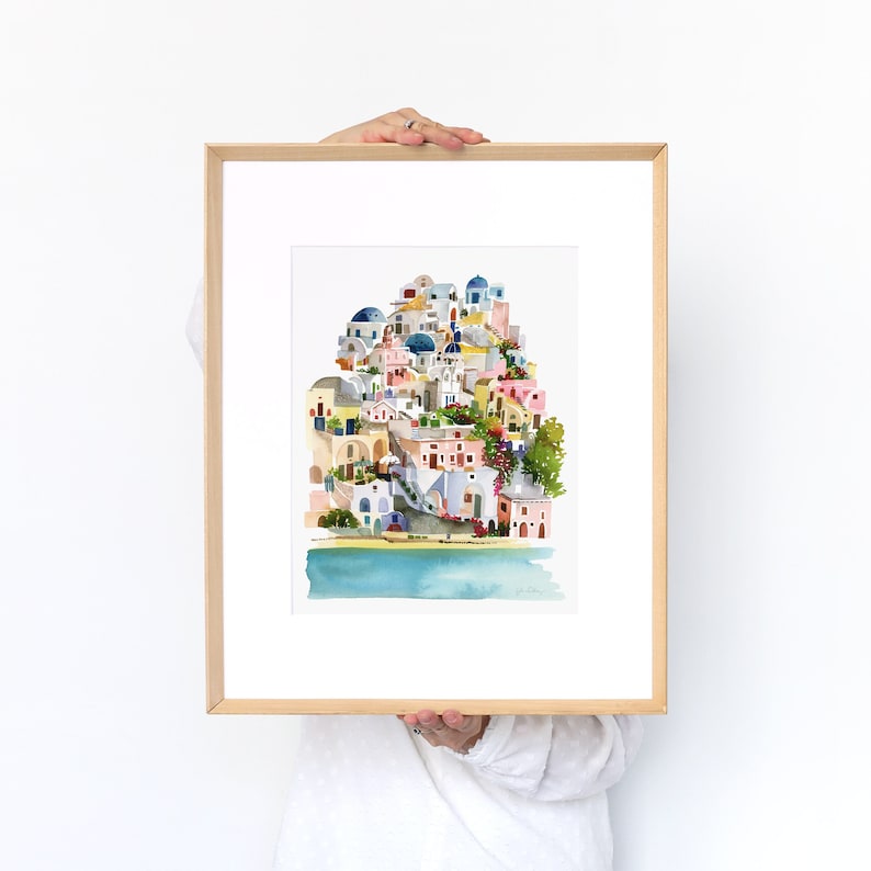 Peut inclure: Une peinture &agrave; l'aquarelle d'un village color&eacute; avec des b&acirc;timents aux d&ocirc;mes bleus, des murs blanchis &agrave; la chaux et une mer bleu turquoise au premier plan. La peinture est encadr&eacute;e dans un cadre en bois clair.