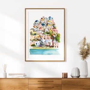 Peut inclure: Une peinture &agrave; l'aquarelle d'un village color&eacute; avec des b&acirc;timents blancs, des d&ocirc;mes bleus et une mer bleu turquoise. Le village est construit &agrave; flanc de colline et les b&acirc;timents sont regroup&eacute;s. La peinture est encadr&eacute;e dans un cadre en bois clair.