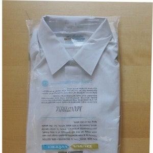 Könnte beinhalten: Ein weißes, langärmeliges Hemd in einer Plastiktüte. Das Hemd ist mit "Manhattan" und "Dura-Wite!" gekennzeichnet und wird als "permanently pressed" beschrieben. Das Etikett gibt an, dass das Hemd aus 85% Polyester und 35% Baumwolle besteht. Das Etikett gibt auch an, dass das Hemd "nie gebügelt werden muss", "leicht zu waschen" ist und "schnell trocknet".