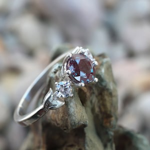 May include: A silver ring featuring a large, oval-cut, color-changing alexandrite gemstone. The ring has a delicate design with a smaller, square-cut stone and leaf-like accents. The alexandrite appears burgundy in the image.