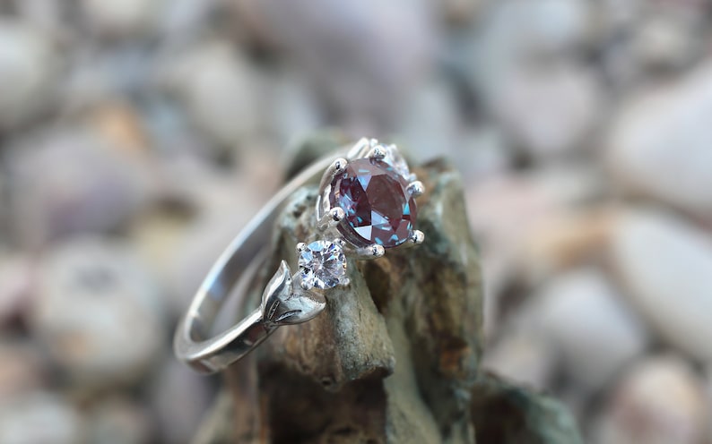 May include: A silver ring featuring a large, oval-cut, color-changing alexandrite gemstone. The ring has a delicate design with a smaller, square-cut stone and leaf-like accents. The alexandrite appears burgundy in the image.
