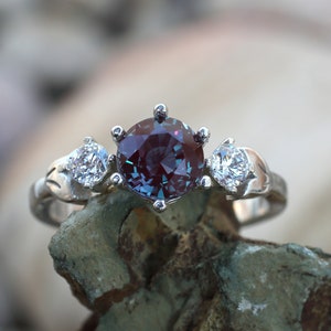 May include: A silver ring with a large, round, color-changing gemstone in the center, flanked by two smaller, clear gemstones. The ring's band has a decorative, leaf-like design. The central stone appears to shift between purple and teal hues.