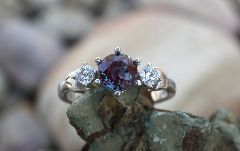 May include: A silver ring with a large, round, color-changing gemstone in the center, flanked by two smaller, clear gemstones. The ring's band has a decorative, leaf-like design. The central stone appears to shift between purple and teal hues.
