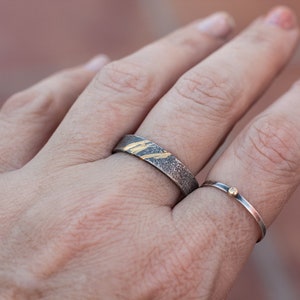 Op de afbeelding: Twee zilveren ringen met gouden accenten. De grotere ring heeft een getextureerde band met dunne gouden lijnen. De kleinere ring heeft een dunne band met een klein gouden stipje.