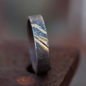 Op de afbeelding: Een zilveren ring met een textuur en gouden accenten. De ring is een eenvoudige band met een diagonaal patroon van gouden lijnen.