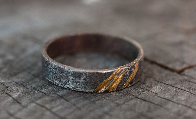 Op de afbeelding: Een zilveren ring met een getextureerde afwerking en gouden accenten. De ring heeft een brede band en een eenvoudig ontwerp.
