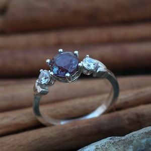 May include: A silver ring featuring a large, round, purplish-blue gemstone flanked by two smaller, clear stones. The ring's band has a simple, elegant design, showcasing the gemstones prominently. The ring is set against a blurred, warm-toned background.