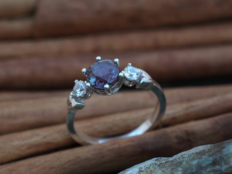 May include: A silver ring featuring a large, round, purplish-blue gemstone flanked by two smaller, clear stones. The ring's band has a simple, elegant design, showcasing the gemstones prominently. The ring is set against a blurred, warm-toned background.