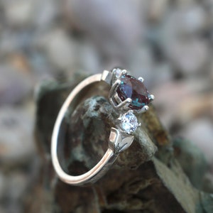 May include: A silver ring featuring a large, oval, color-changing alexandrite gemstone and a smaller, round, clear gemstone. The ring has a delicate, open design with leaf accents. The gemstones are set against a blurred background of rocks.