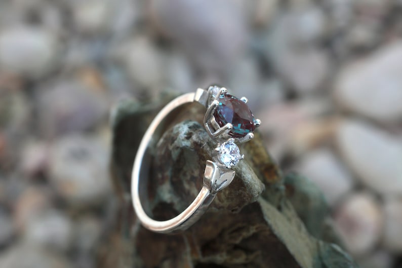 May include: A silver ring featuring a large, oval, color-changing alexandrite gemstone and a smaller, round, clear gemstone. The ring has a delicate, open design with leaf accents. The gemstones are set against a blurred background of rocks.