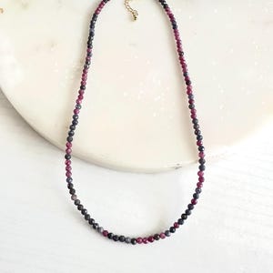 May include: A delicate necklace featuring small, round beads in shades of deep red, dark blue, and gray. The necklace has a gold-colored clasp and chain. The beads are strung together to create a simple, elegant design.