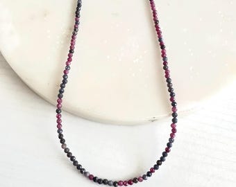 May include: A delicate necklace featuring small, round beads in shades of deep red, dark blue, and gray. The necklace has a gold-colored clasp and chain. The beads are strung together to create a simple, elegant design.