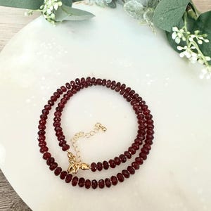 May include: A double-strand necklace featuring small, round, deep red beads. The necklace is secured with a gold-colored clasp and extender chain. The jewelry is displayed on a white surface with greenery in the background.