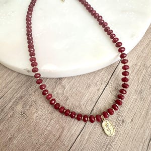 May include: A burgundy beaded necklace with a gold-colored clasp and a small pendant. The necklace features small, round beads and a delicate gold-colored pendant with a floral design.
