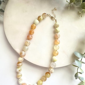 May include: A beaded necklace featuring round, faceted stones in a gradient of warm colors including pale yellow, soft orange, cream, and light green. The beads are strung on a gold-toned wire with a matching gold-toned metal clasp and extender chain.