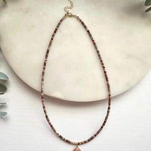 May include: A beaded necklace featuring small, multi-colored earth-toned stones in shades of brown, tan, and grey. The necklace includes a gold-toned metal clasp with an extender chain and a single, smooth, teardrop-shaped peach-colored pendant.