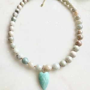 May include: A beaded necklace featuring a strand of round, neutral-toned beads. A turquoise heart-shaped pendant hangs from the center. The necklace has a gold-colored clasp and extender chain. The beads are approximately 0.8 cm in diameter.