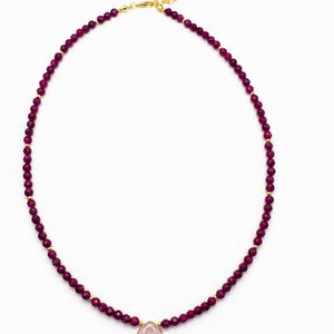 May include: A necklace featuring a strand of small, faceted deep magenta beads interspersed with tiny gold-toned spacers. A single, smooth, pale pink teardrop-shaped pendant hangs from the center. The necklace includes a gold-toned metal clasp.