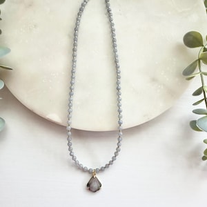 May include: A delicate necklace featuring small, light gray beads strung together with a gold-colored clasp. A seashell pendant in shades of gray and gold hangs from the center. The necklace is displayed on a white marble surface.