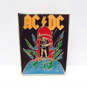 May include: A black and gold enamel pin featuring the band AC/DC. The pin depicts a figure standing on a globe with a rocket behind them. The band's name is written in yellow letters with a lightning bolt.