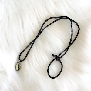May include: A black cord necklace with a small oval pendant featuring a yin-yang symbol. The pendant has a silver-colored border and a yellow and black design. The necklace is displayed on a white, fluffy surface.