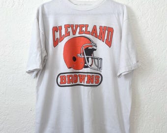 May include: White t-shirt featuring the words "CLEVELAND BROWNS" in orange block letters above and below a football helmet graphic. The helmet is orange with a silver facemask. The shirt is hanging on a black hanger.