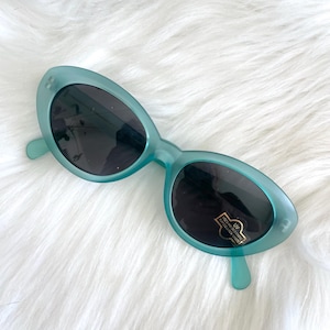 May include: Teal-colored oval sunglasses with dark lenses. The sunglasses have a retro design and are set against a white, fluffy background. A small sticker is on the lens.