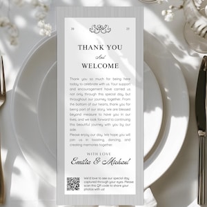 Könnte beinhalten: Eine Hochzeitstafel mit einer silbernen Gabel, einem Messer und einem Löffel. Ein weißer Teller trägt eine graue Karte mit dem Text "THANK YOU AND WELCOME" und den Namen "Emilia & Michael". Die Karte enthält einen QR-Code.