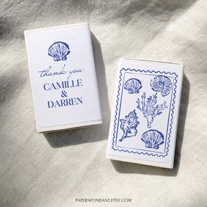 May include: Two white matchboxes with blue seashell designs. One box has the text "thank you CAMILLE & DARREN". The other box features a blue seashell and coral design within a blue scalloped border. The boxes are on a white linen surface.
