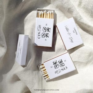 May include: Several white matchboxes with black text and illustrations. The text includes "CIN Cin", "20", and "CHEERS TO Allie & Greg". The matches have wooden stems and white tips. The boxes are on a light-colored fabric.