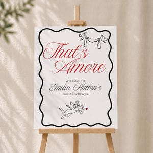 May include: A white rectangular sign with a black wavy border featuring the red text That's Amore, black line art of two birds holding a banner, and a cupid figure. Below, black text reads Welcome to Emilia Hutton's Bridal Shower.