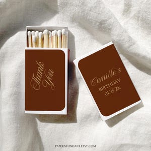 Può includere: Vengono mostrate due scatole di fiammiferi marroni con bordi bianchi. Una scatola è aperta, rivelando fiammiferi con punte bianche. La prima scatola recita "Thank You" in corsivo dorato. La seconda scatola recita "Camille's Birthday 01.25.2X" in corsivo dorato.
