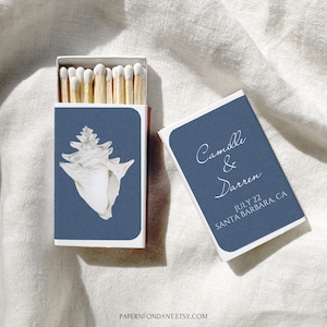 May include: Matchbox with a navy blue cover featuring a white seashell illustration. The open matchbox reveals wooden matches with white tips. A separate navy blue matchbox cover displays the names "Camille & Darren" and the date "July 22 SANTA BARBARA, CA."